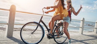 two women riding bikes on a boardwalk near the ocean.